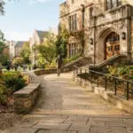 A wide, gently sloping, sun-drenched stone accessibility ramp leading to a stately university building entrance